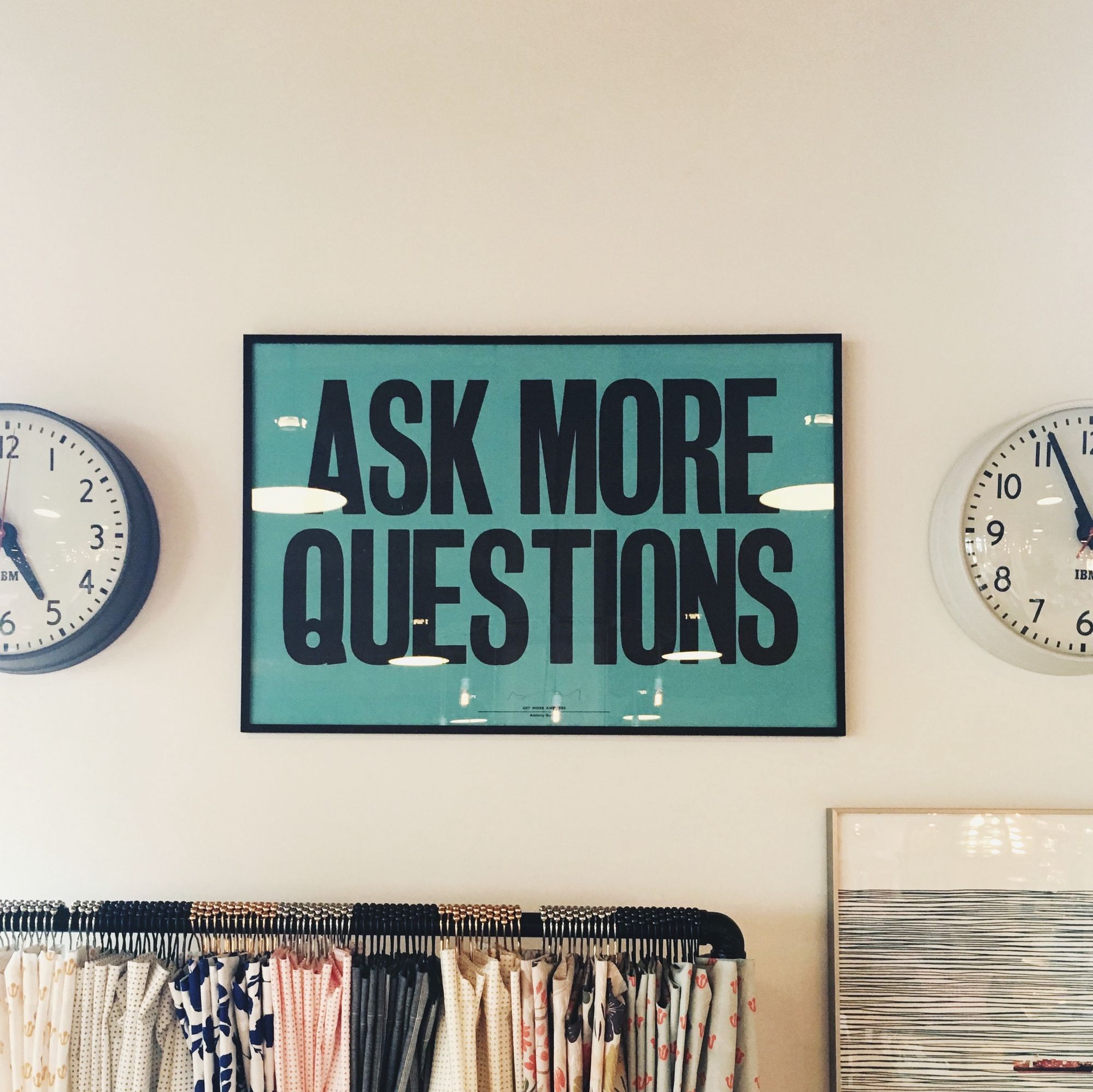 A teal colored graphic poster with the text "Ask More Questions" hanging on a beige wall between two IBM branded clocks